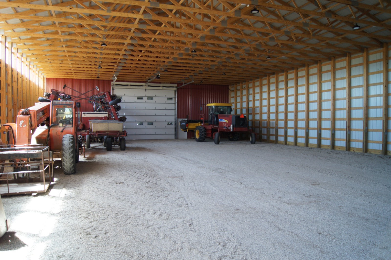 A telehandler, a planter and a swather parked inside a post-frame building with open floor between them.