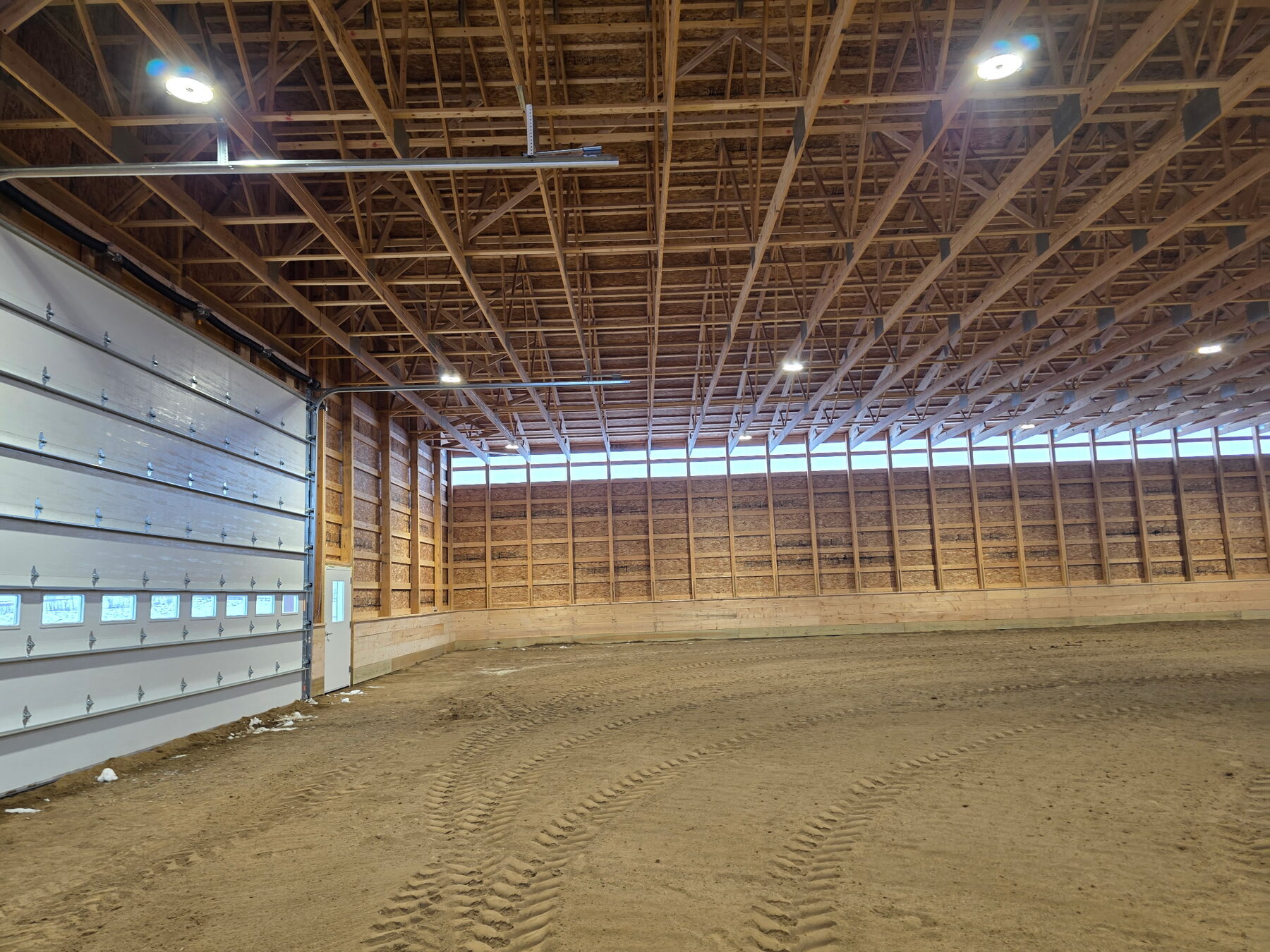 Inside a finished Prairie Barns riding arena: clear span timber trusses, lighting down the ridge, sand footing and a full-height door at the end.